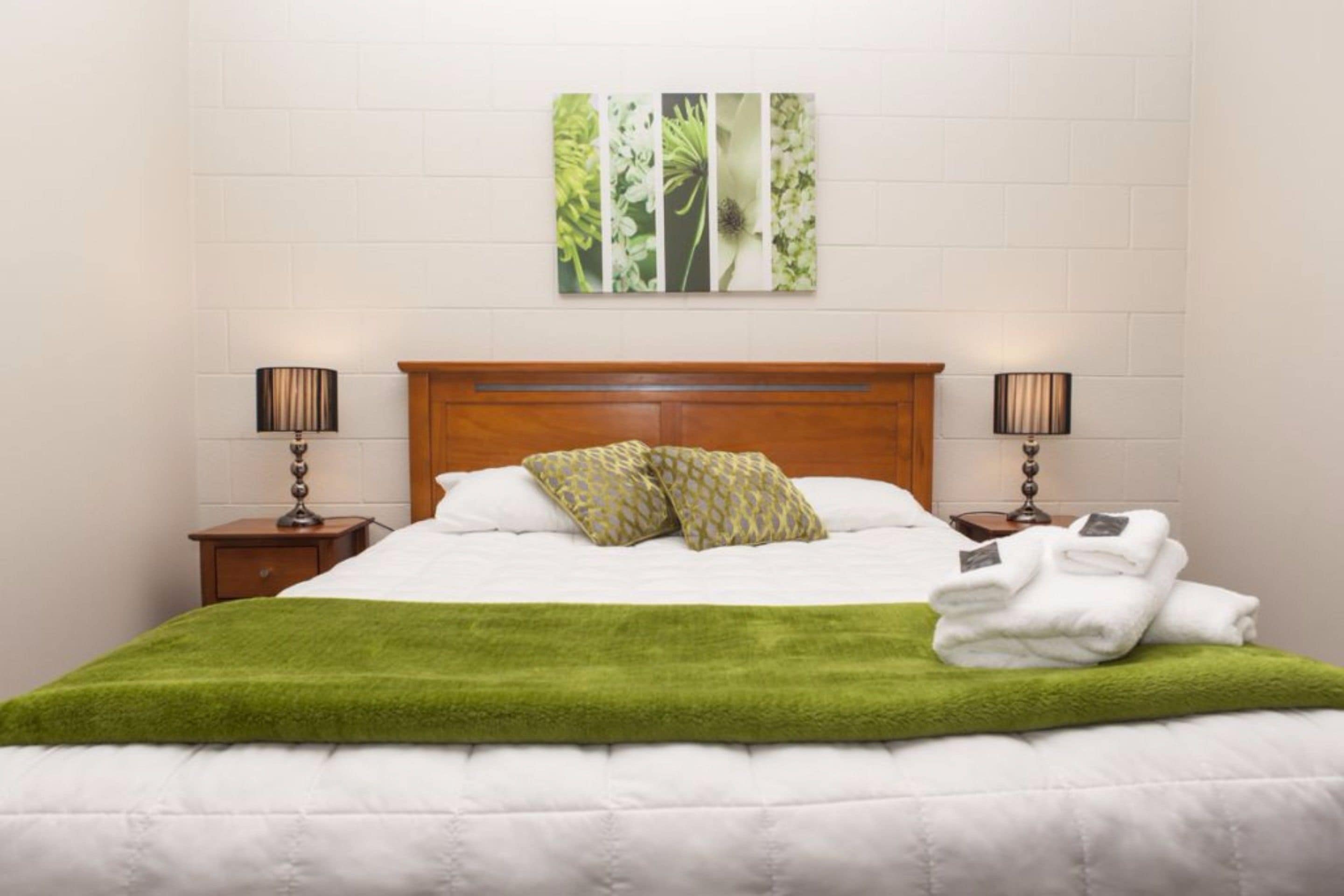 A neatly made bed with a green throw blanket, two bedside lamps, a wooden headboard, and a floral wall art piece above the bed.