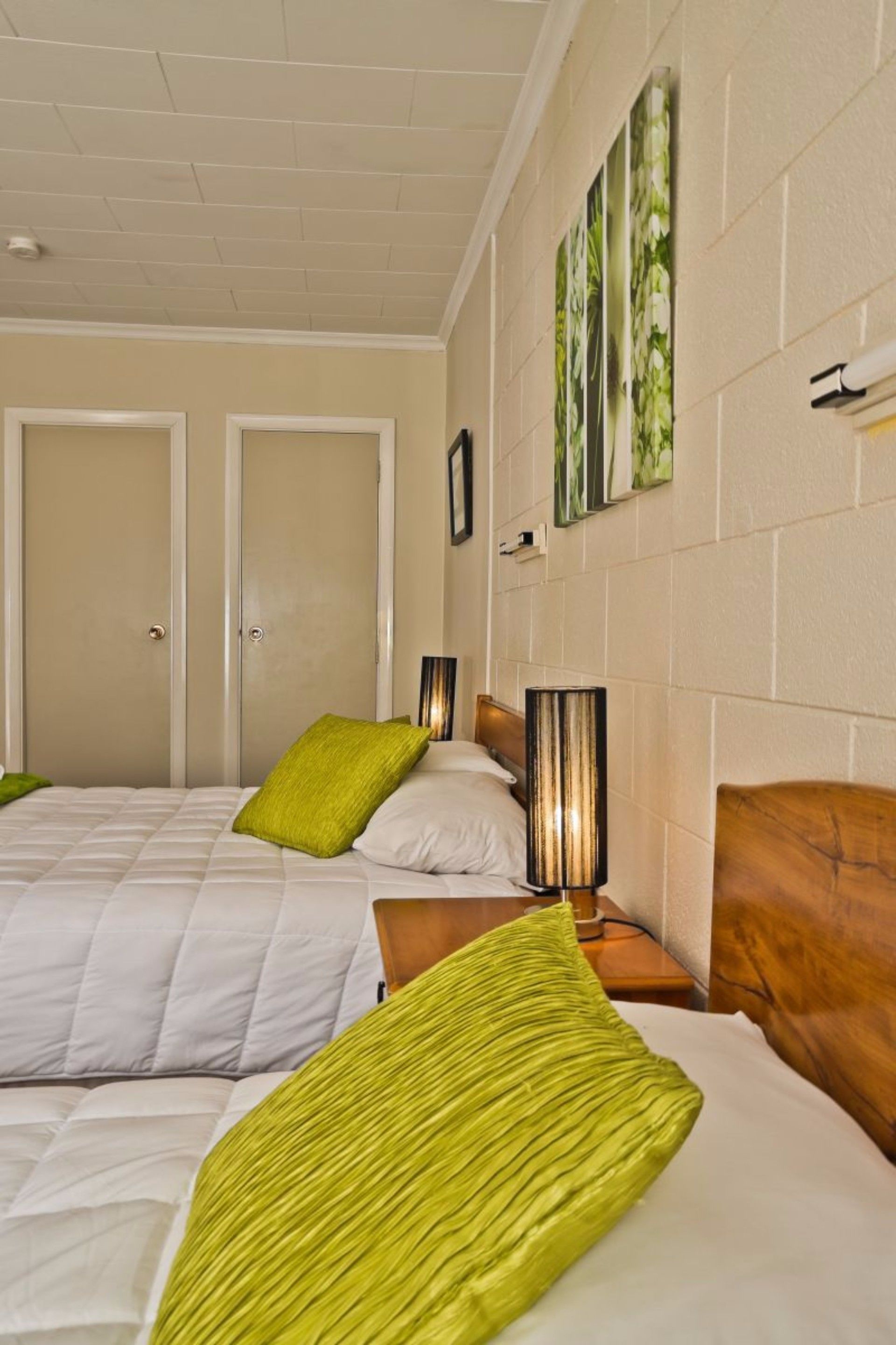 Quarto de hotel com duas camas, cada uma com cabeceira de madeira, roupa de cama branca e almofadas verdes. Há luminárias fixadas na parede em ambos os lados das camas.