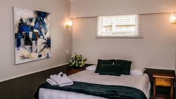Een keurig opgemaakte slaapkamer met een bed, een nachtkastje, een kunstwerk aan de muur en een raam met zonwering.