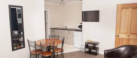 A compact room with a dining table, chairs, a sofa, a TV mounted on the wall, and a kitchenette.