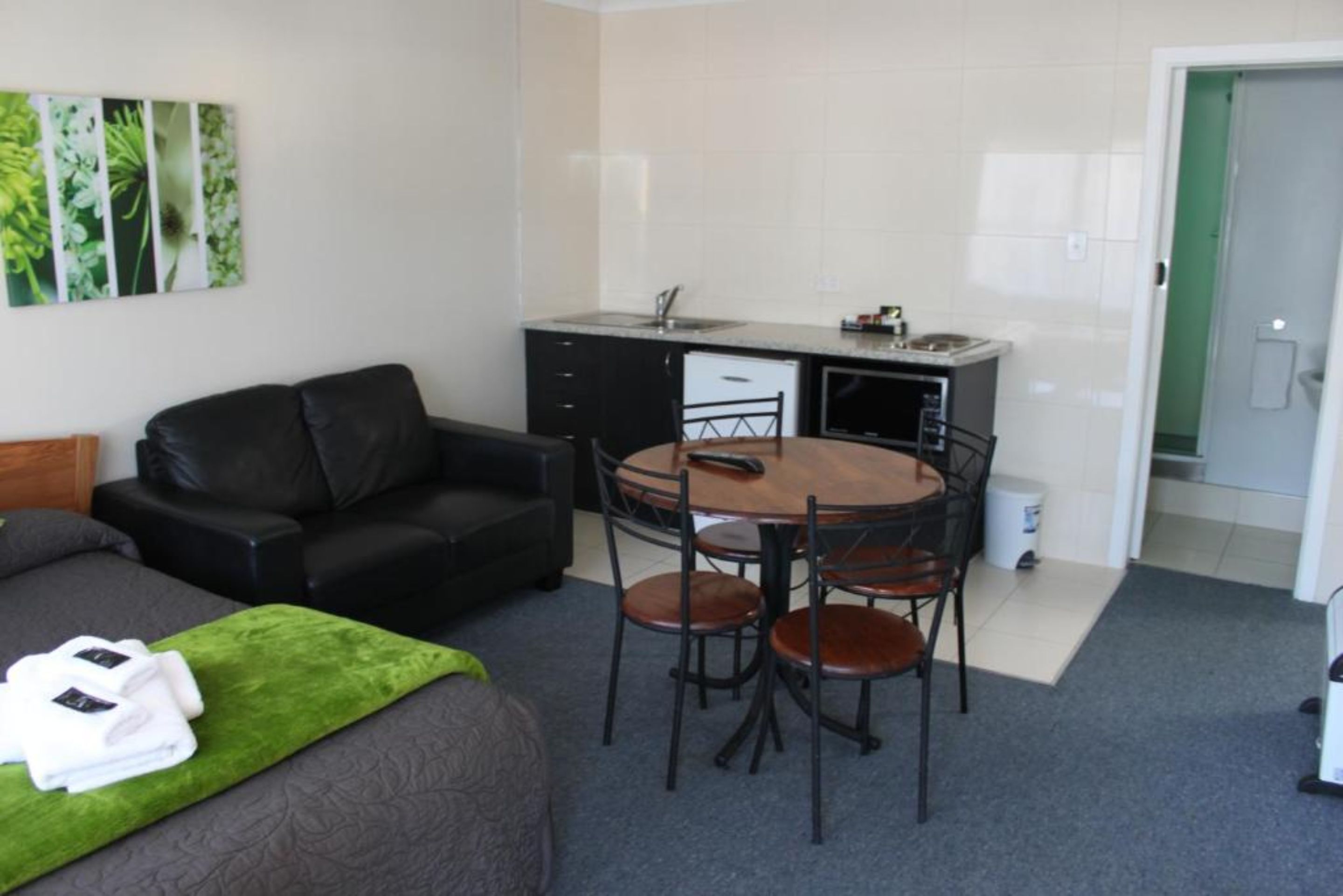 Habitación de hotel compacta con cama, alfombra verde, sofá de cuero negro, pequeña mesa de comedor con cuatro sillas, zona de cocina con fregadero y microondas, y una puerta que da a un baño.