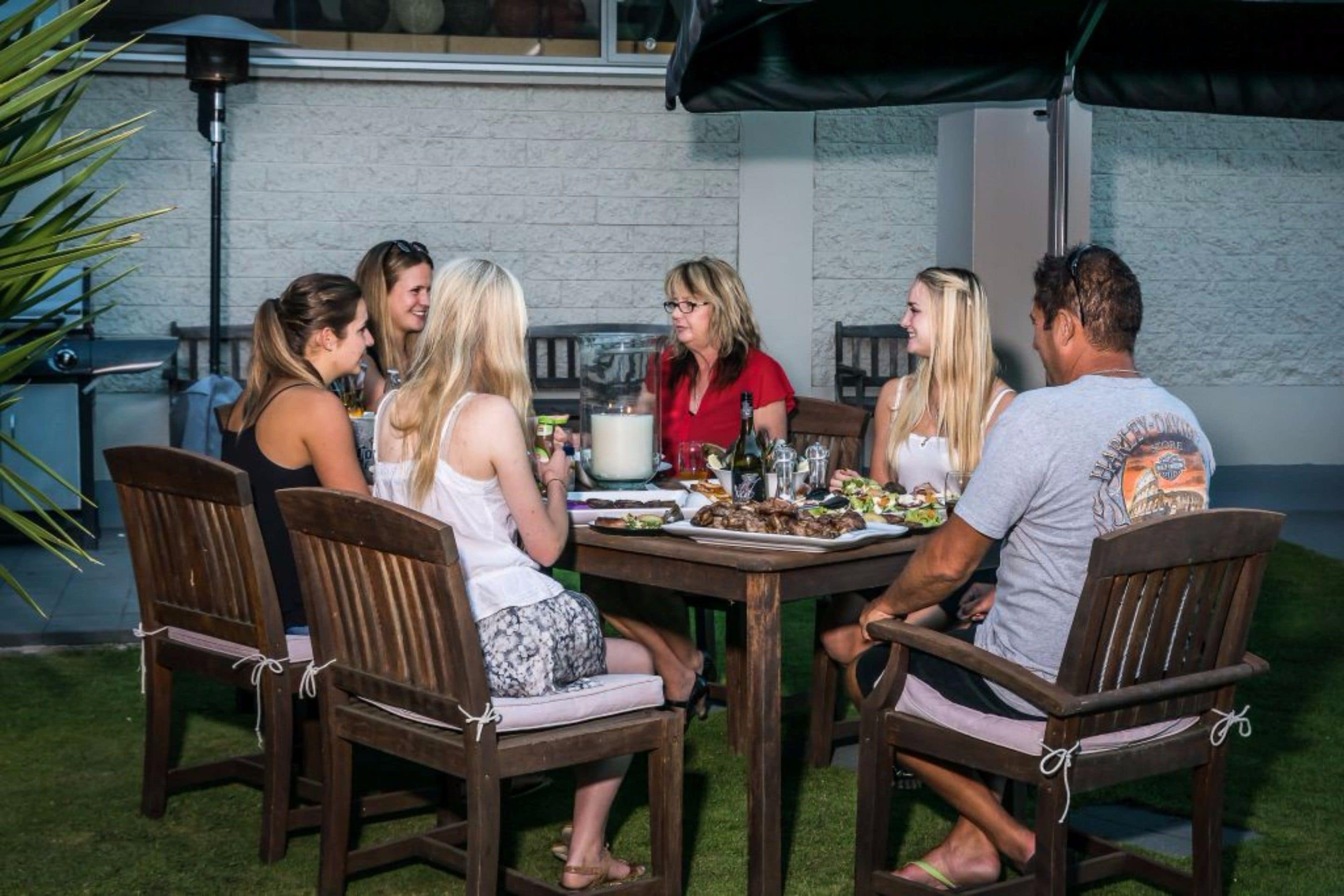 Un grupo de personas sentadas alrededor de una mesa de madera al aire libre, con un muro de ladrillo al fondo.
