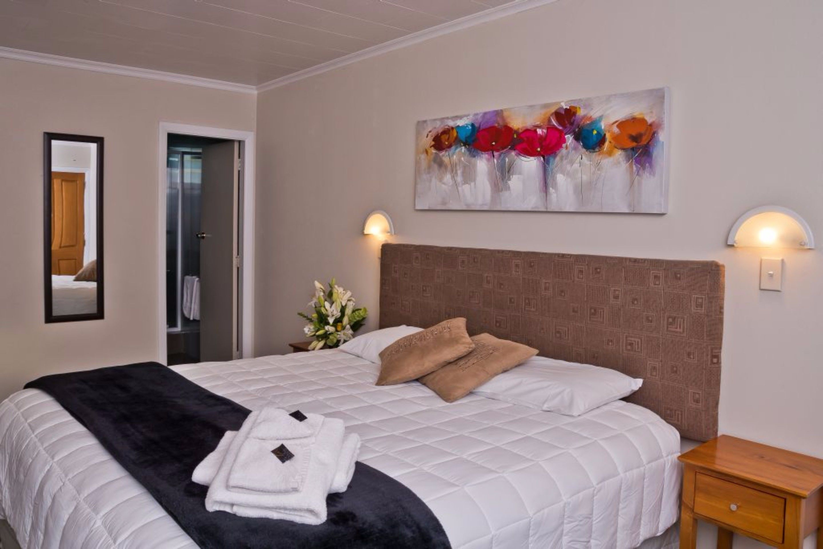 A neatly made bed with a white quilt, two pillows, and folded towels. There is a wall-mounted painting of flowers and two wall sconces.