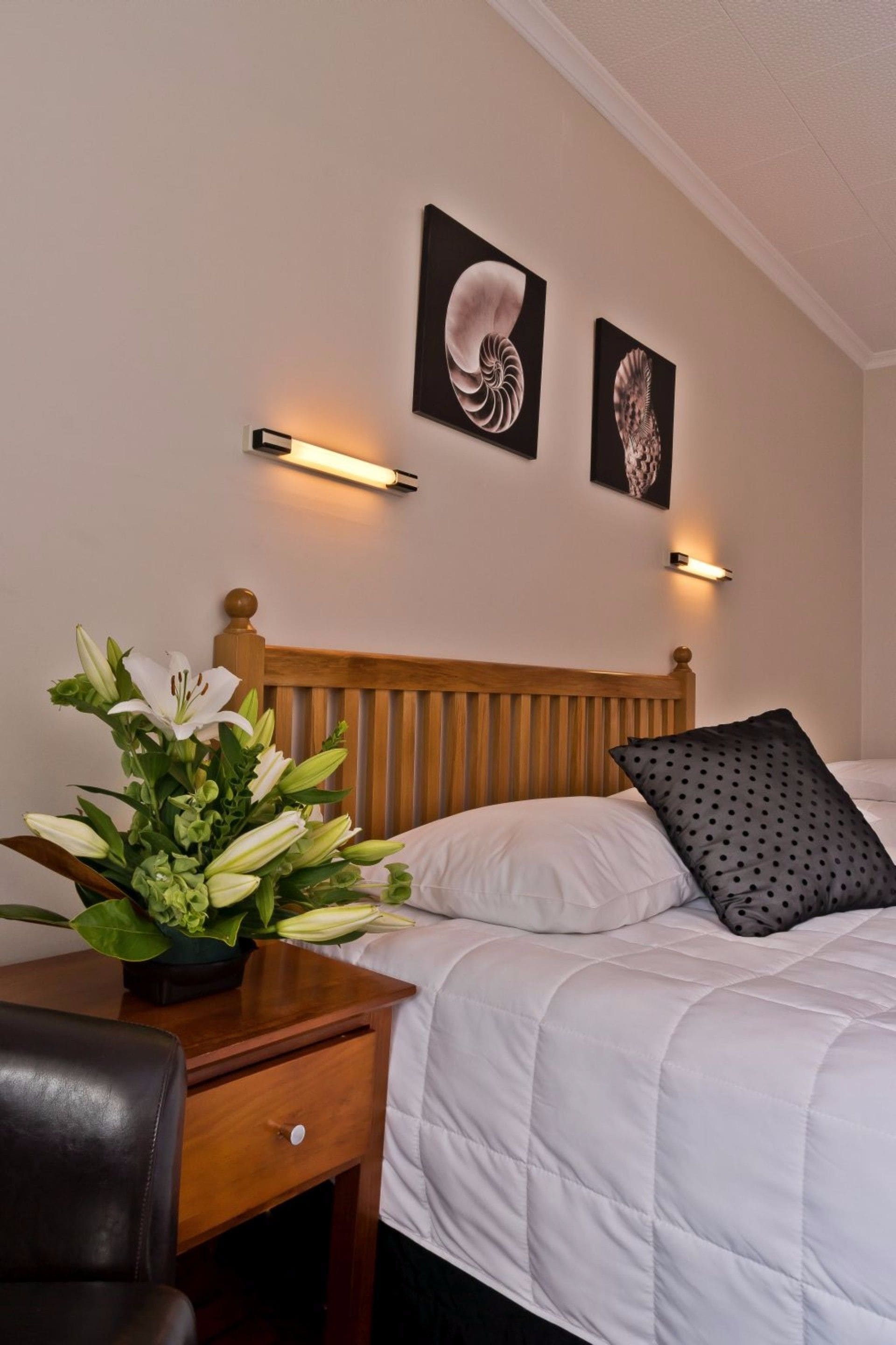 Habitación de hotel con cabecera de madera, ropa de cama blanca, un cojín de lunares negros, una mesita de noche con un florero y dos lámparas fijadas a la pared.