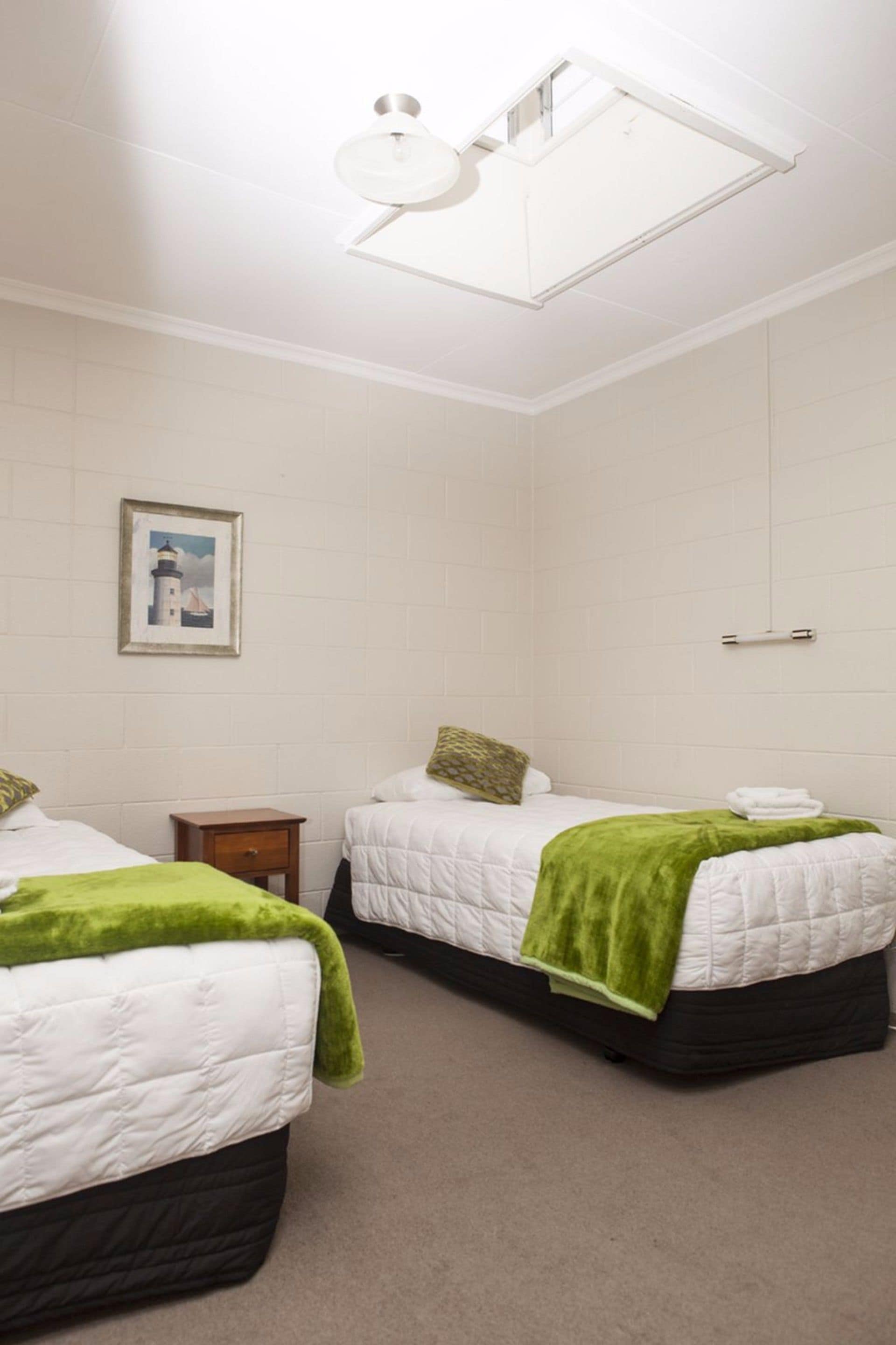 A room with two beds, a small bedside table, a wall-mounted light fixture, and a framed picture on the wall.