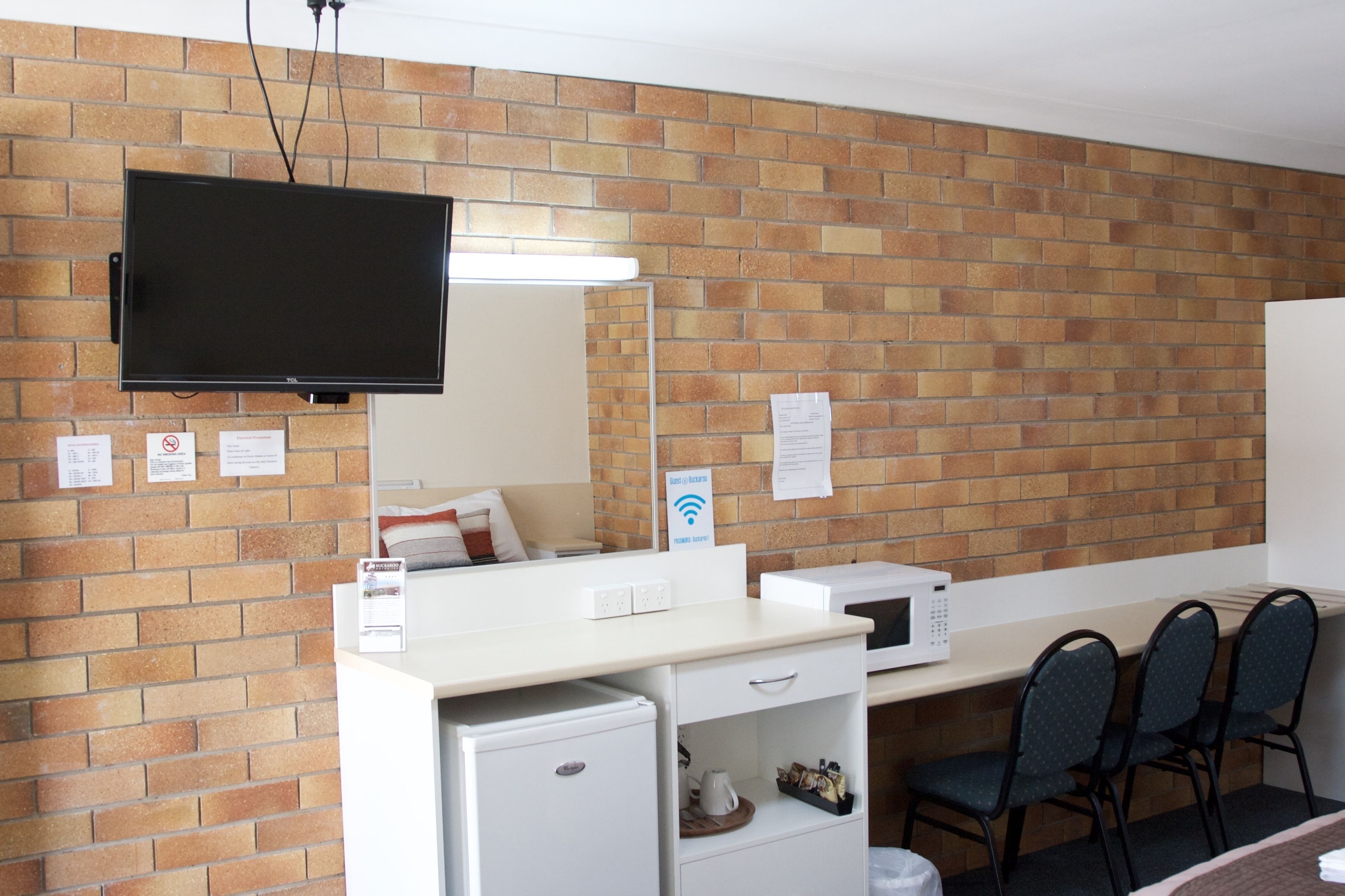 A hotel room with a brick wall, a flat-screen TV mounted above a desk, a microwave, a mini-fridge, and a seating area with chairs.