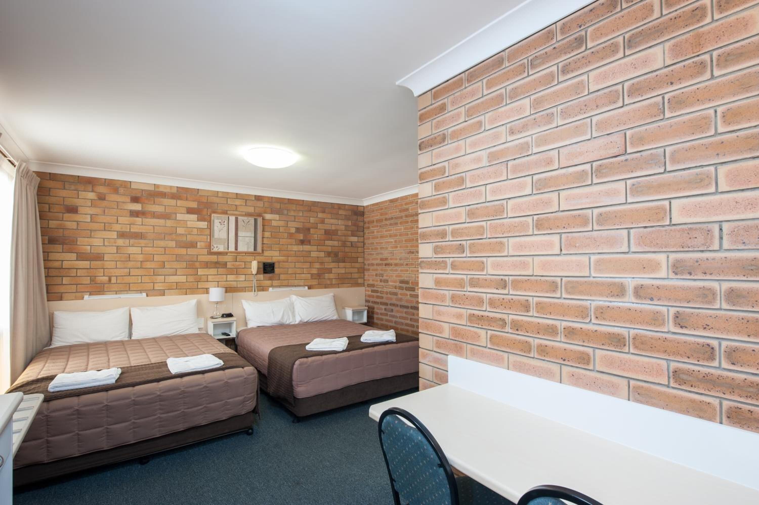 A hotel room with two beds, a desk, and a brick wall.