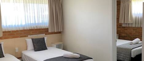 A hotel room with two single beds, a small bedside table, a headboard with a brick accent, and a window with curtains.