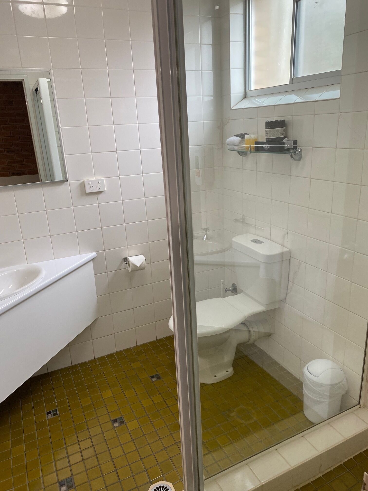 A bathroom with a white sink, toilet, and shower area. There is a mirror above the sink and a shelf with toiletries.
