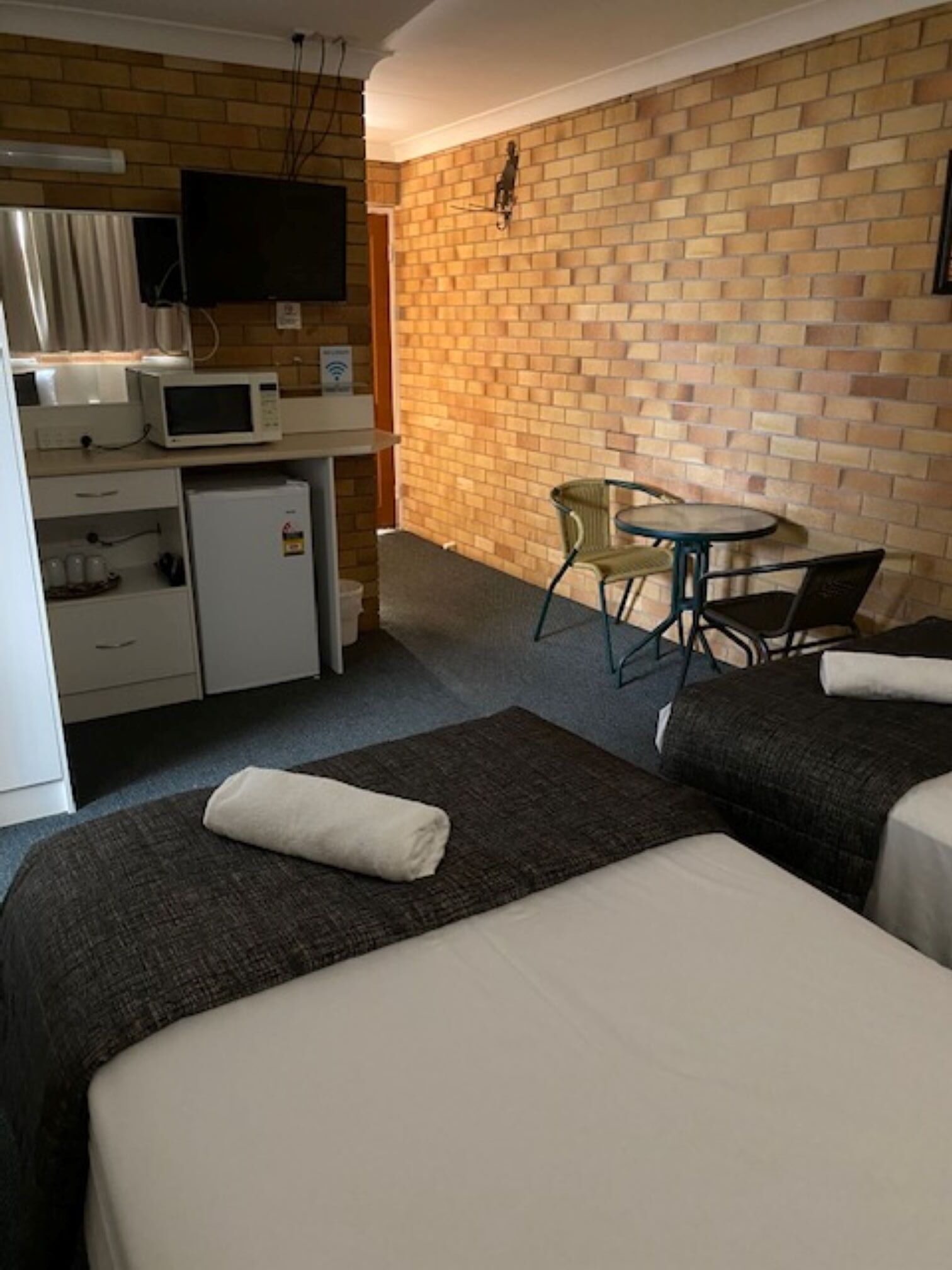 Habitación de hotel con dos camas, una kitchenette pequeña, un televisor montado en la pared y una pared con diseño de ladrillos.