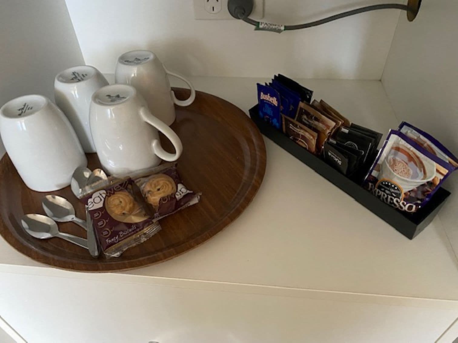Una encimera de cocina con tazas blancas, una bandeja de madera con dos cucharas y una caja de galletas, y una estantería con paquetes de café y una caja de bizcochos.