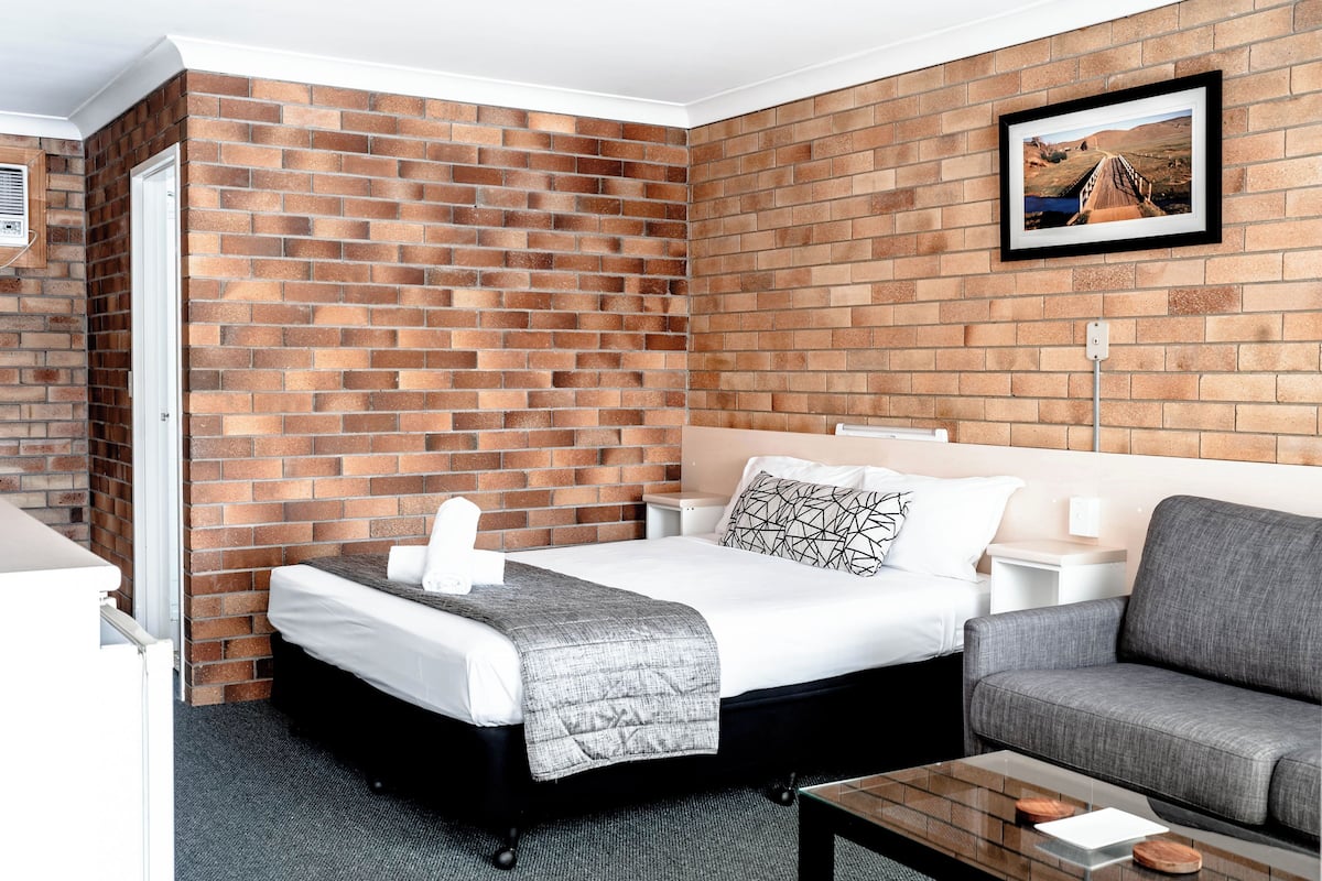 Habitación de hotel con pared de ladrillo, cama con cubrecama blanco y manta gris, sofá gris y un cuadro en la pared.