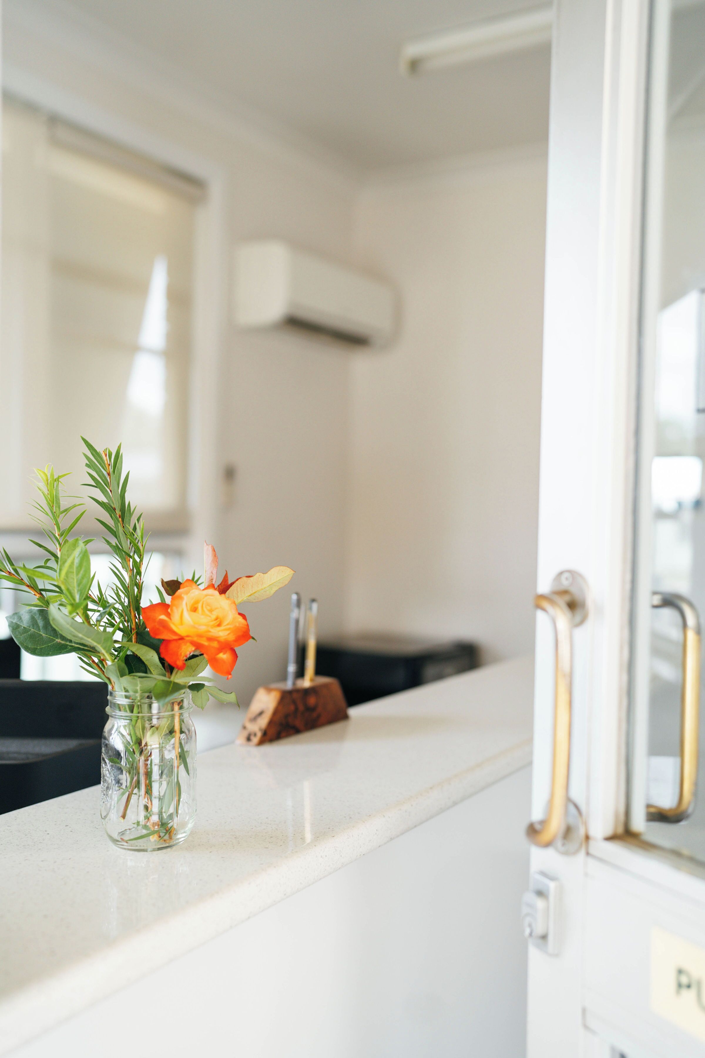 A bathroom with a white countertop, a glass jar with a plant and a single orange flower, a wooden object, and a mirror with a gold handle.