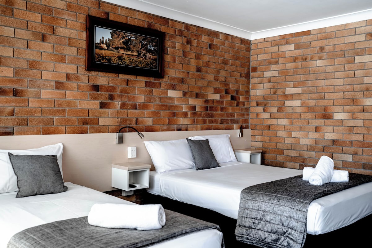 Habitación de hotel con dos camas, una pared de ladrillo, un cuadro enmarcado y una mesita de noche con una lámpara.