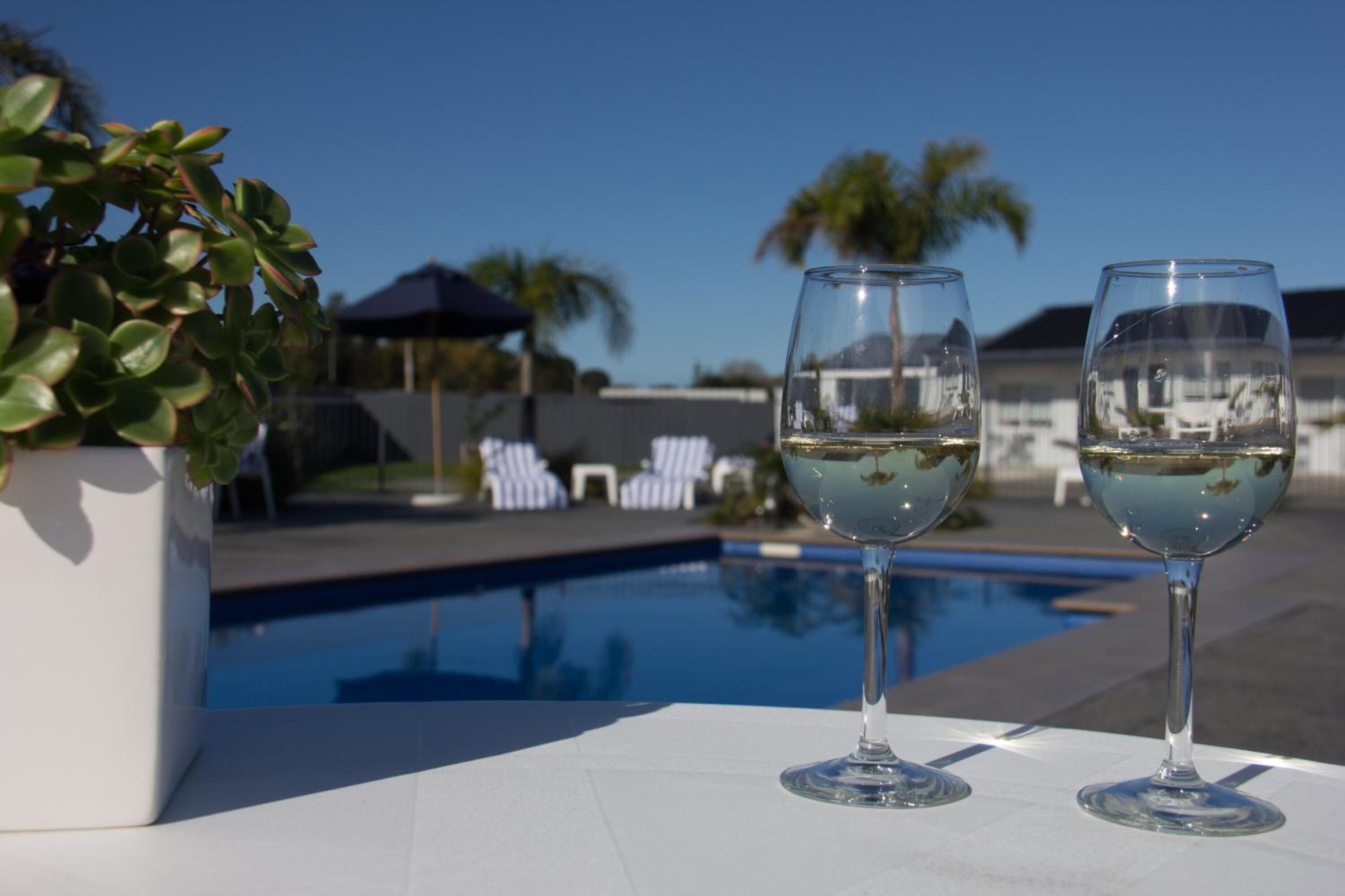 Two glasses of white wine on a poolside table with a clear view of a swimming pool and lounge chairs in the background.