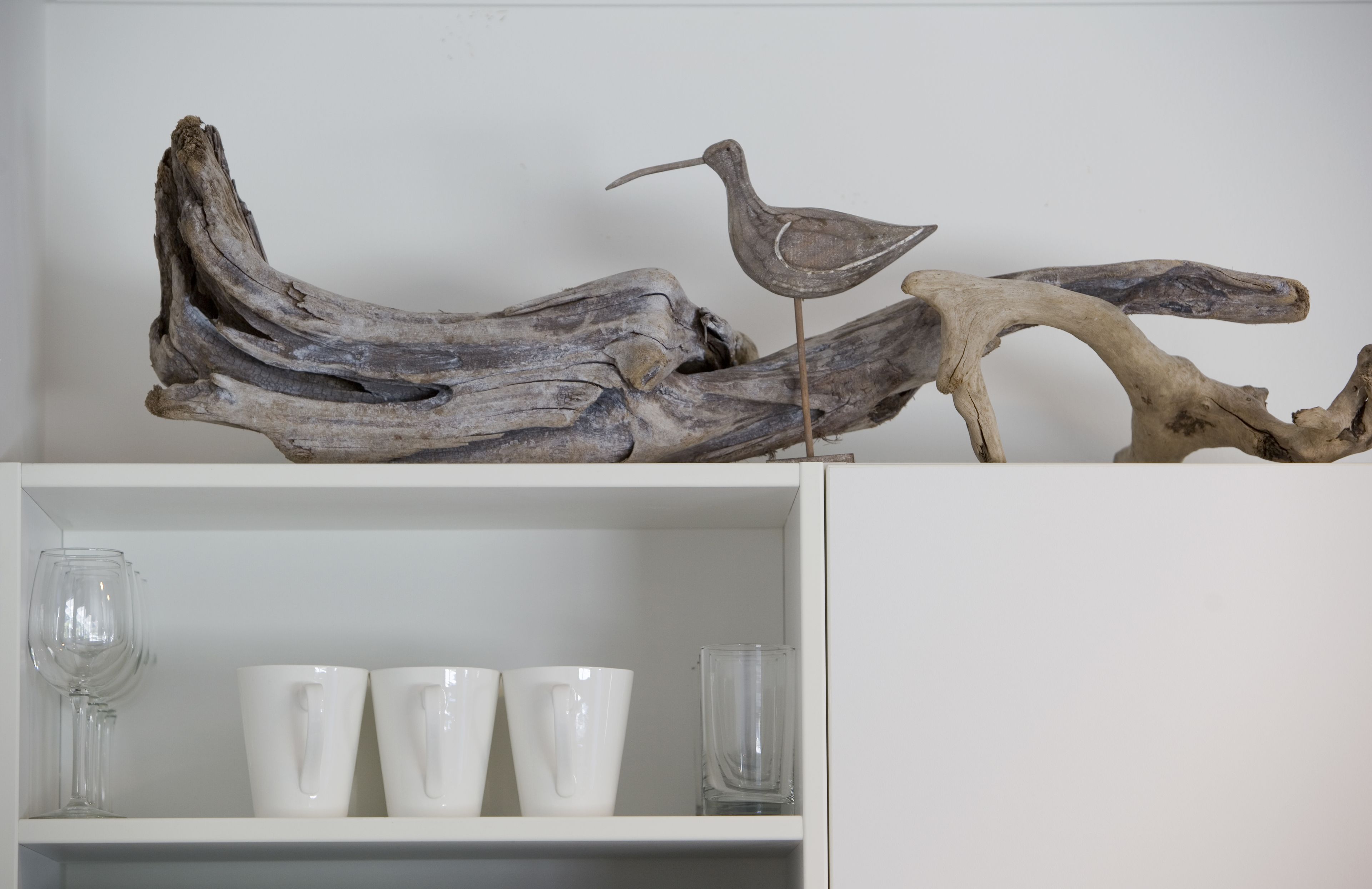 Una repisa con tres tazas blancas y un vaso, además de un trozo de madera a la deriva con una escultura de un pájaro.
