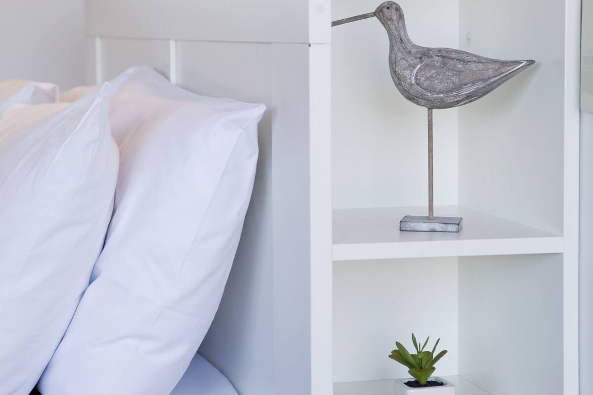 Un dormitorio con una cama blanca, una mesita de noche blanca, una escultura decorativa de un pájaro, una pequeña planta en maceta y una revista sobre la mesita de noche.