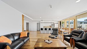 A modern living room with a black leather sofa, a wooden coffee table, and a dining area with a kitchen in the background. - Jenatt Apartments Salamanca (Battery Point)
