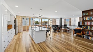 A modern kitchen with a central island, a dining area with chairs, a black leather sofa, and a bookshelf filled with books. - Jenatt Apartments Salamanca (Battery Point)
