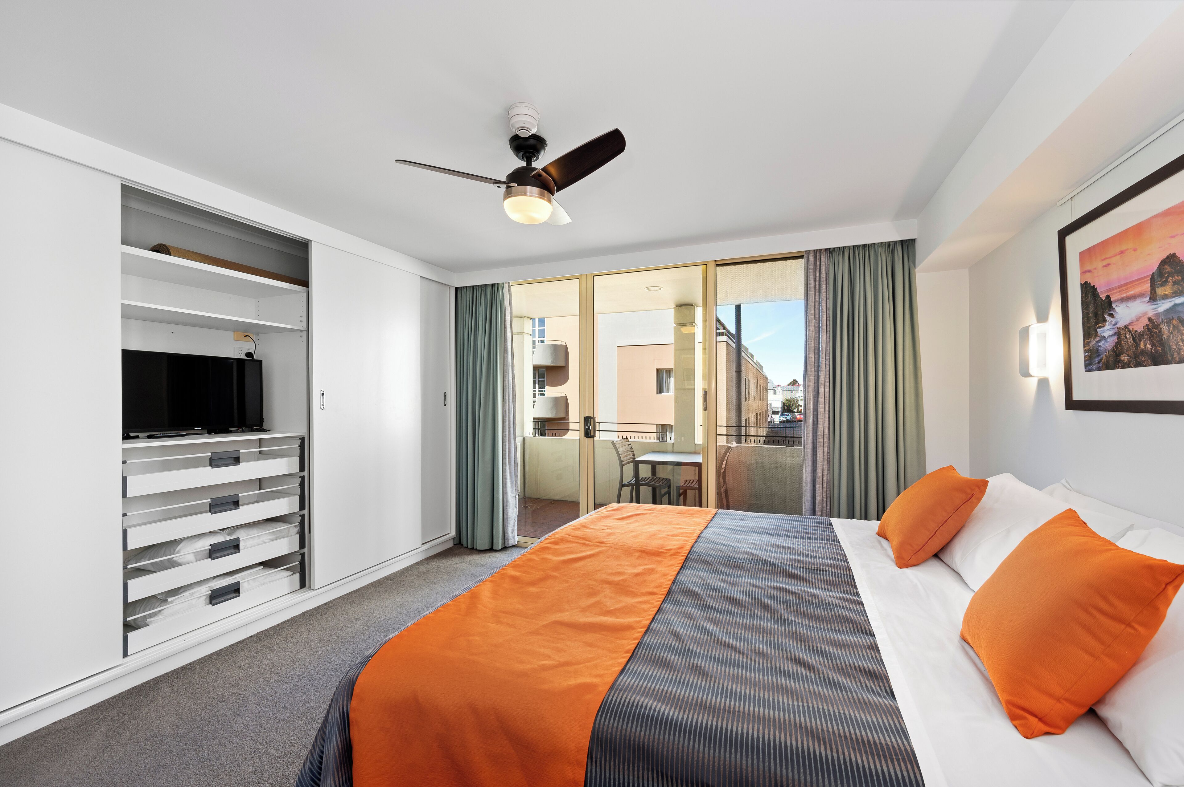 A hotel room with a bed, orange bedding, a TV, a ceiling fan, a balcony with a table and chairs, and a view of buildings.