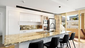 A modern kitchen with granite countertops, white cabinetry, and a large island with bar stools. - Jenatt Apartments Salamanca (Battery Point)