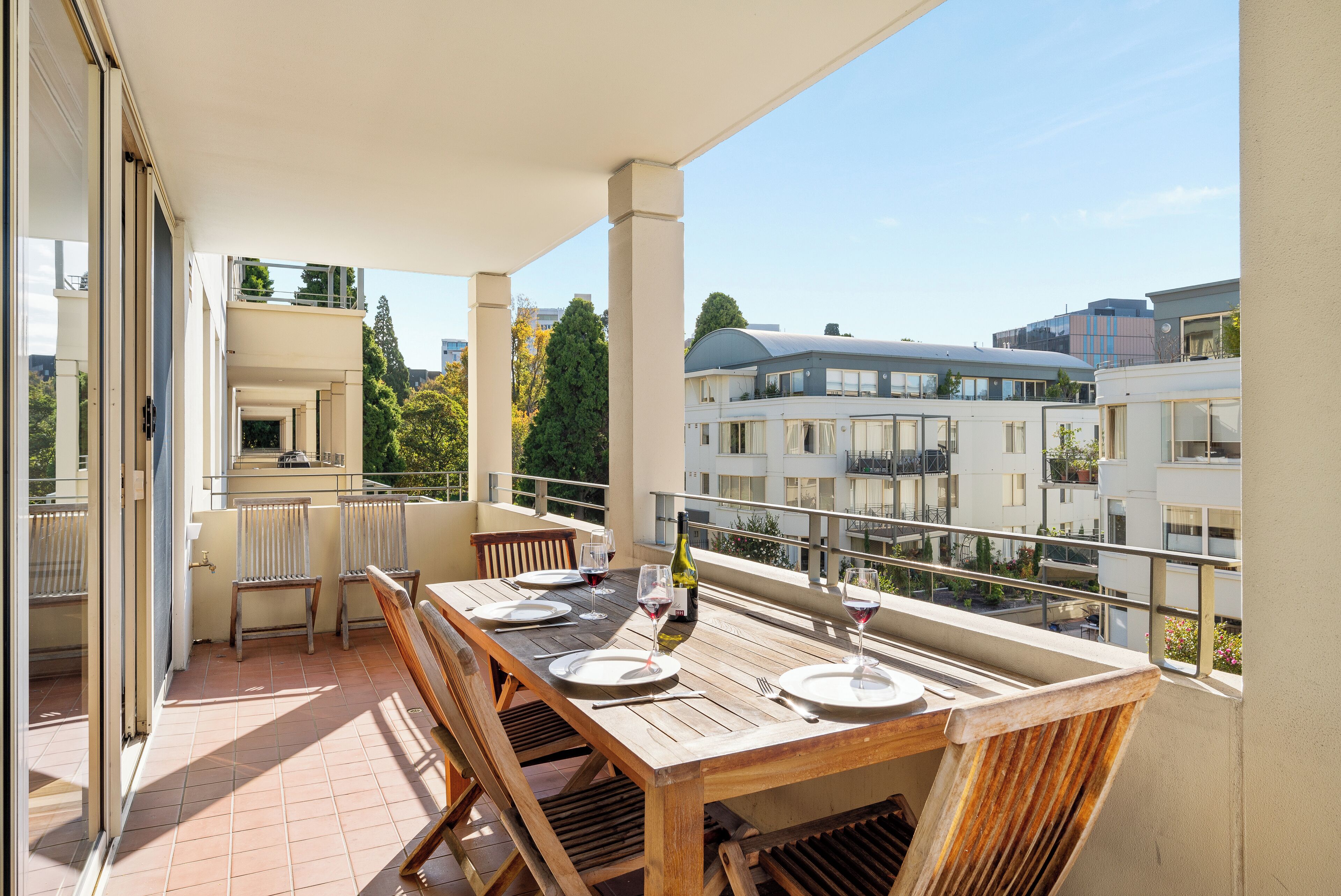 A balcony with a wooden table set for dining, chairs, and wine glasses.