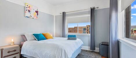 A bedroom with a large bed, a nightstand with a lamp, a window with curtains, and a city view.