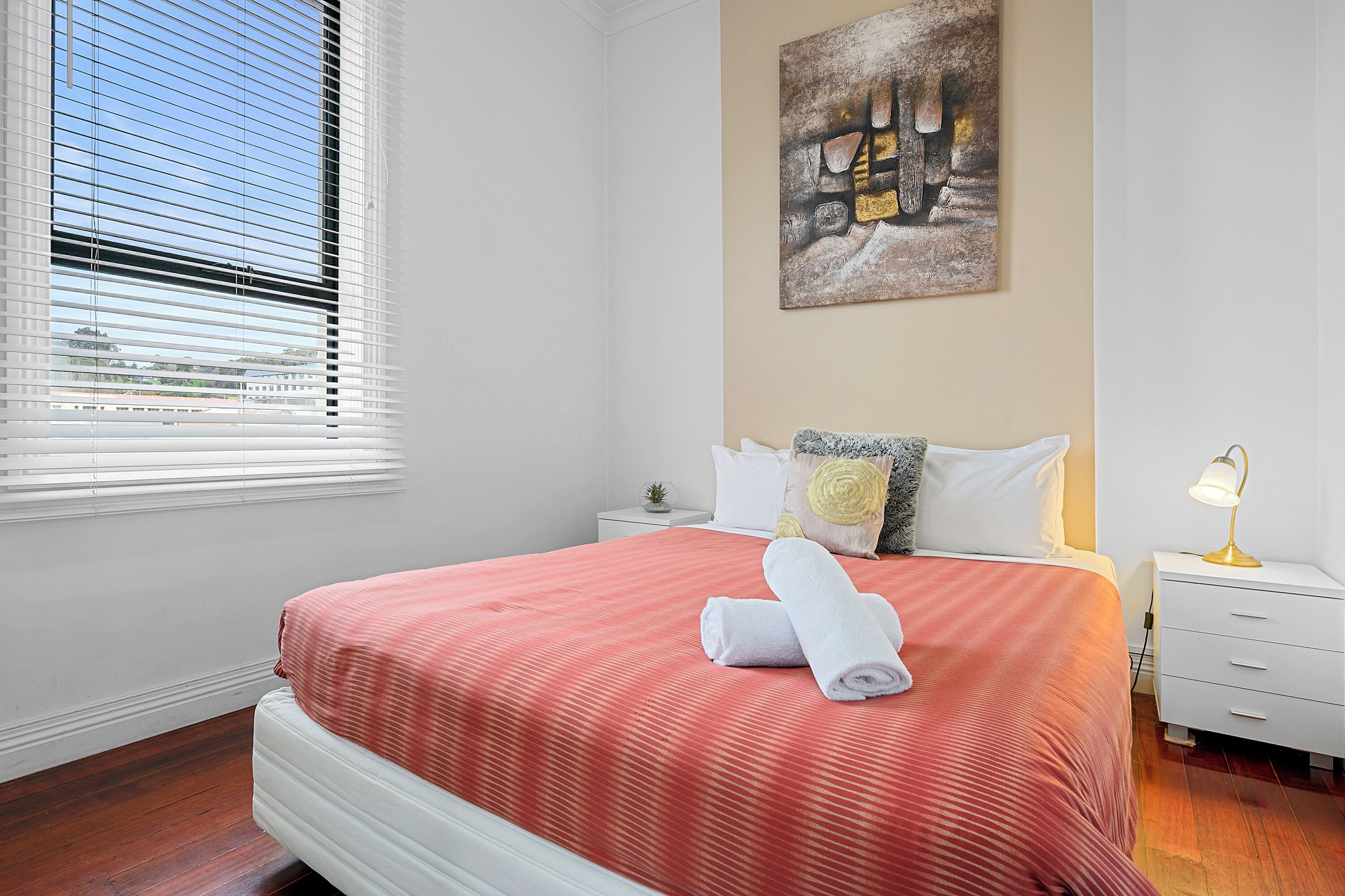 A neatly made bed with a red bedspread and white pillows, a small bedside table with a lamp, and a window with blinds.