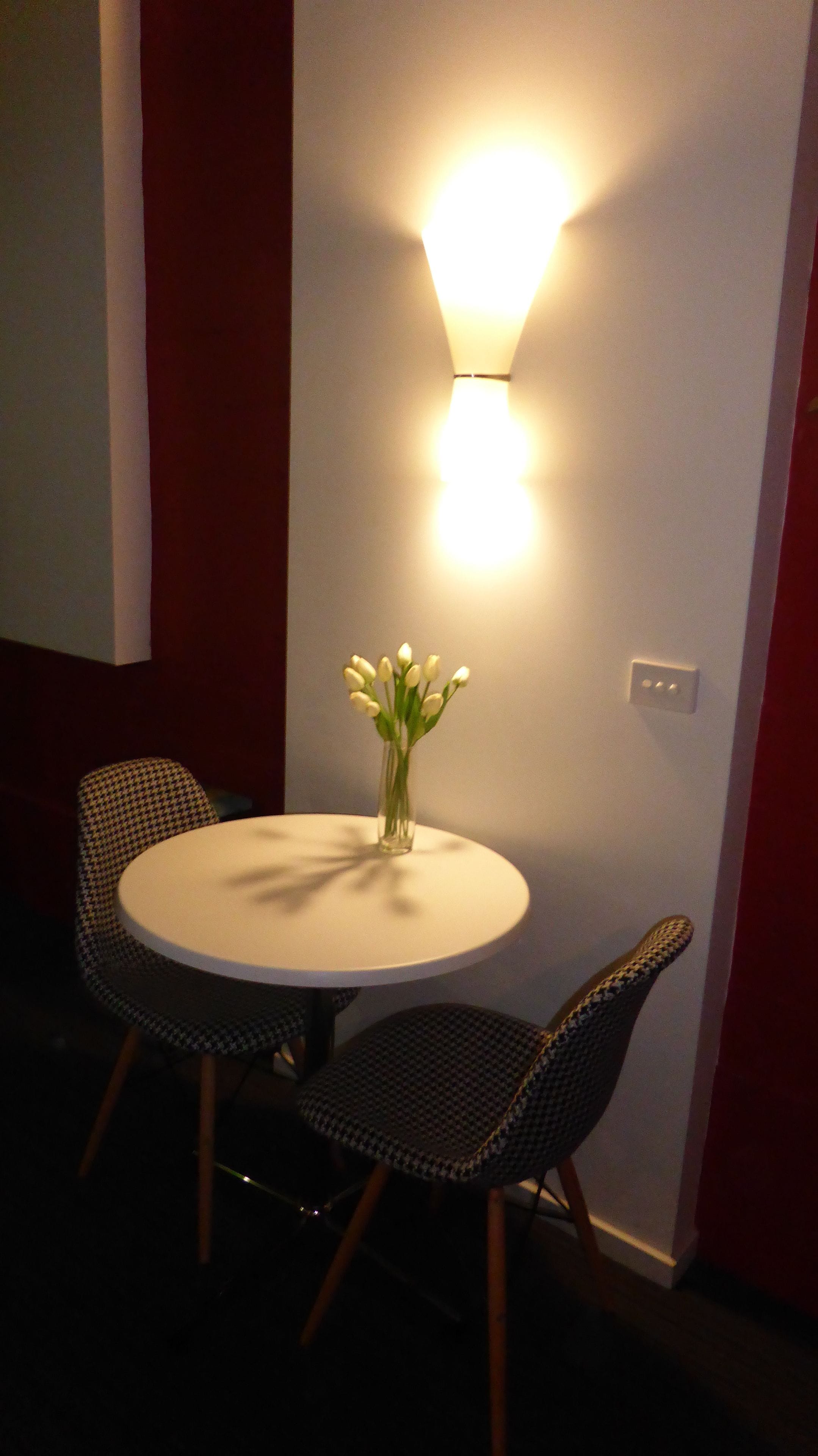 Une table ronde avec deux chaises, un vase rempli de fleurs blanches et un luminaire fixé au mur.