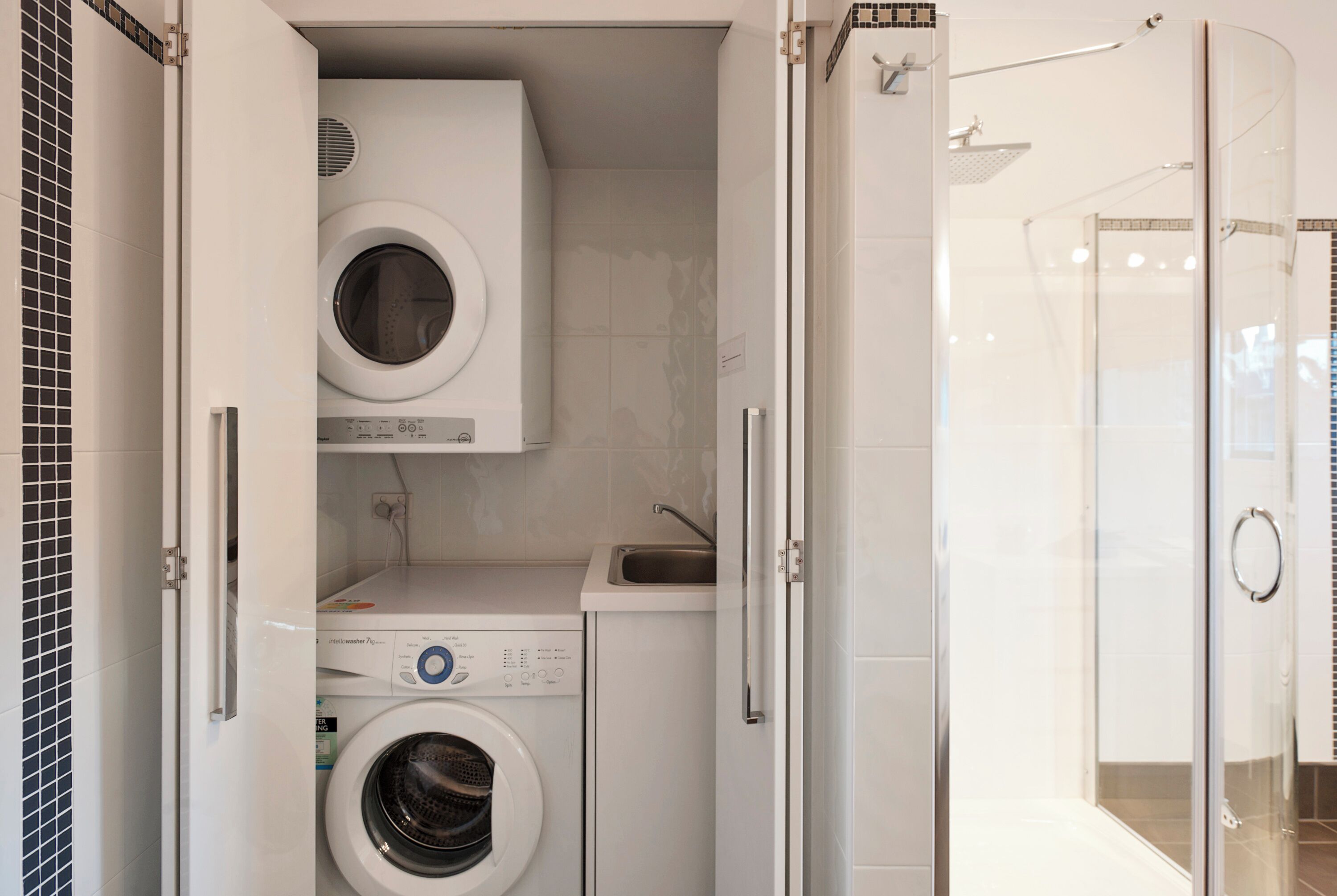 A compact laundry room with a washing machine, dryer, and a glass-enclosed shower area.