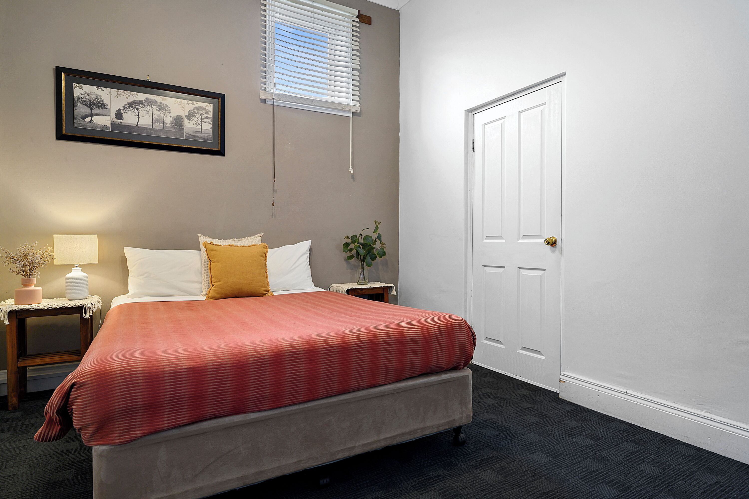 Une chambre à coucher comprenant un lit, des tables de chevet, une lampe, un tableau encadré, une porte et une fenêtre avec des stores.