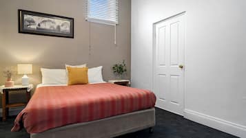 Une chambre à coucher comprenant un lit, des tables de chevet, une lampe, un tableau encadré, une porte et une fenêtre avec des stores.