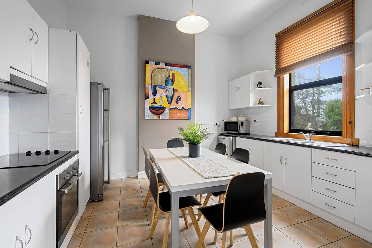 Een moderne keuken met witte kasten, een eettafel met stoelen, een koelkast, een oven en een raam met zonwering.