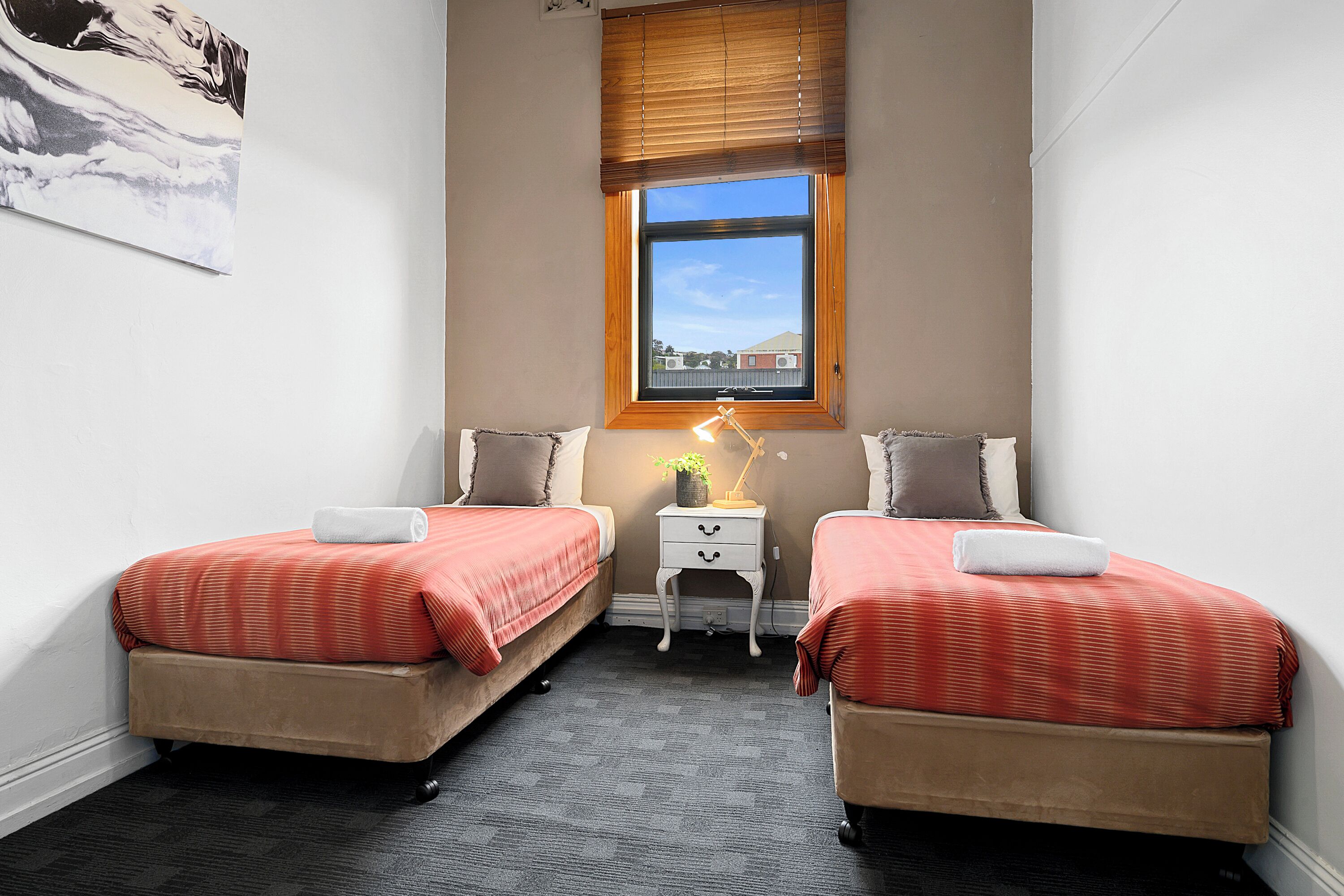 Un dormitorio con dos camas, una mesita de noche, una lámpara y un ventanal con vistas a edificios.