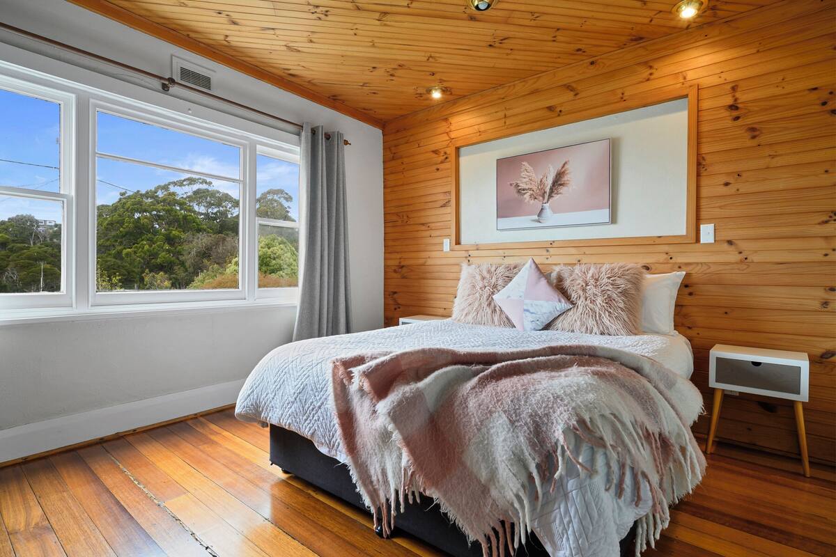 Een slaapkamer met een houten plafond, een groot raam, een bed met een roze deken en een ingelijst kunstwerk aan de muur.