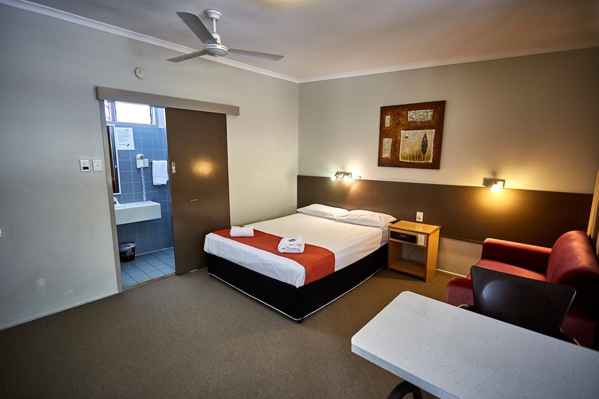 Habitación de hotel con cama, escritorio, silla, ventilador de techo, luz instalada en la pared y puerta que da a un baño.