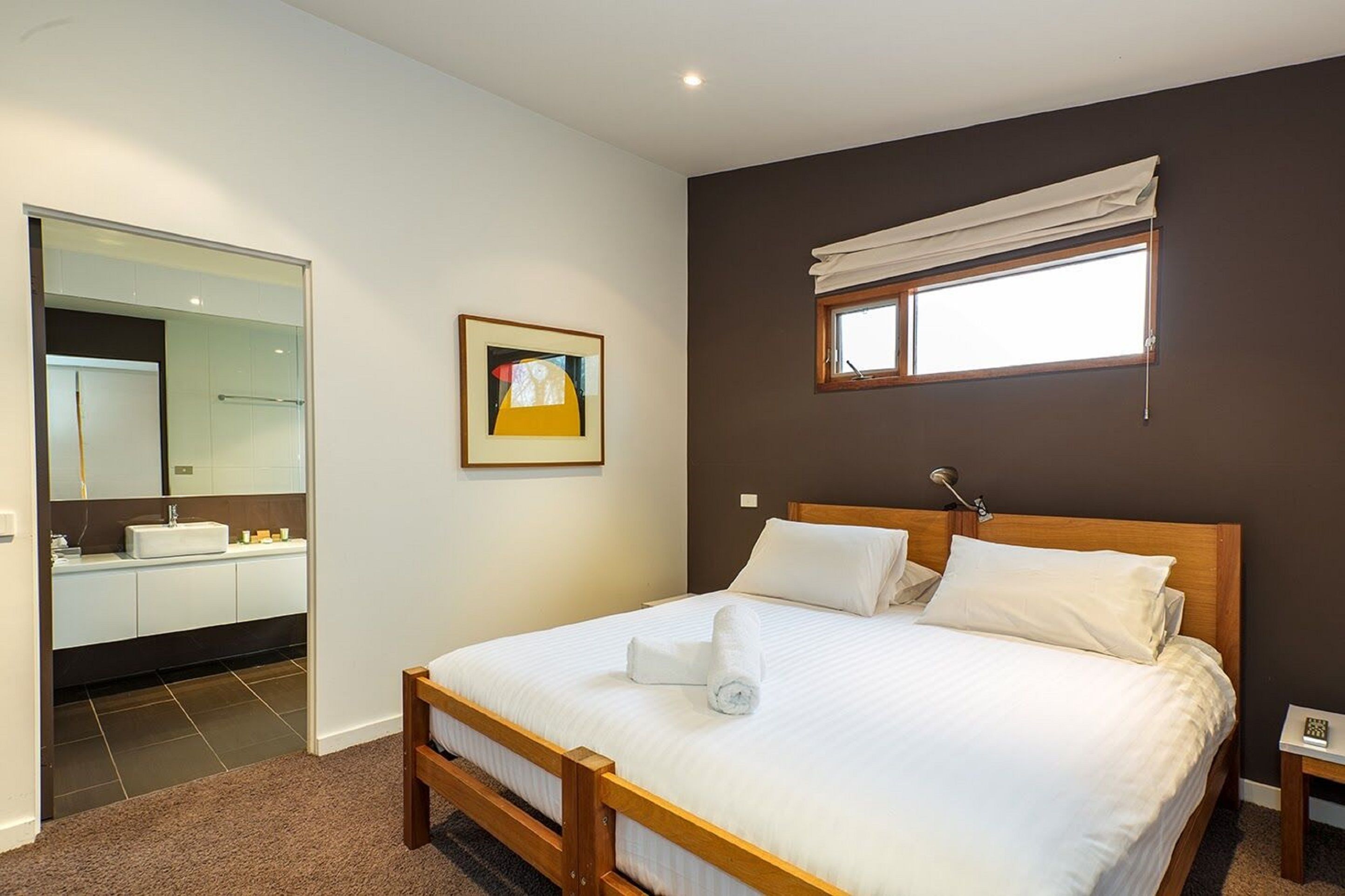 Ein Schlafzimmer mit einem hölzernen Bett, weißer Bettwäsche, einem großen Spiegel, einem gerahmten Bild und einem Fenster mit einem weißen Rollo.