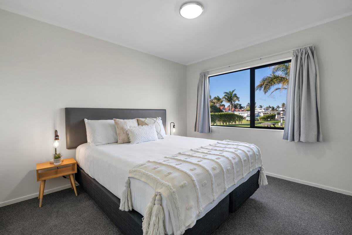 Un dormitorio con una cama grande, una mesita de noche, una ventana con cortinas y vistas a palmeras y casas.