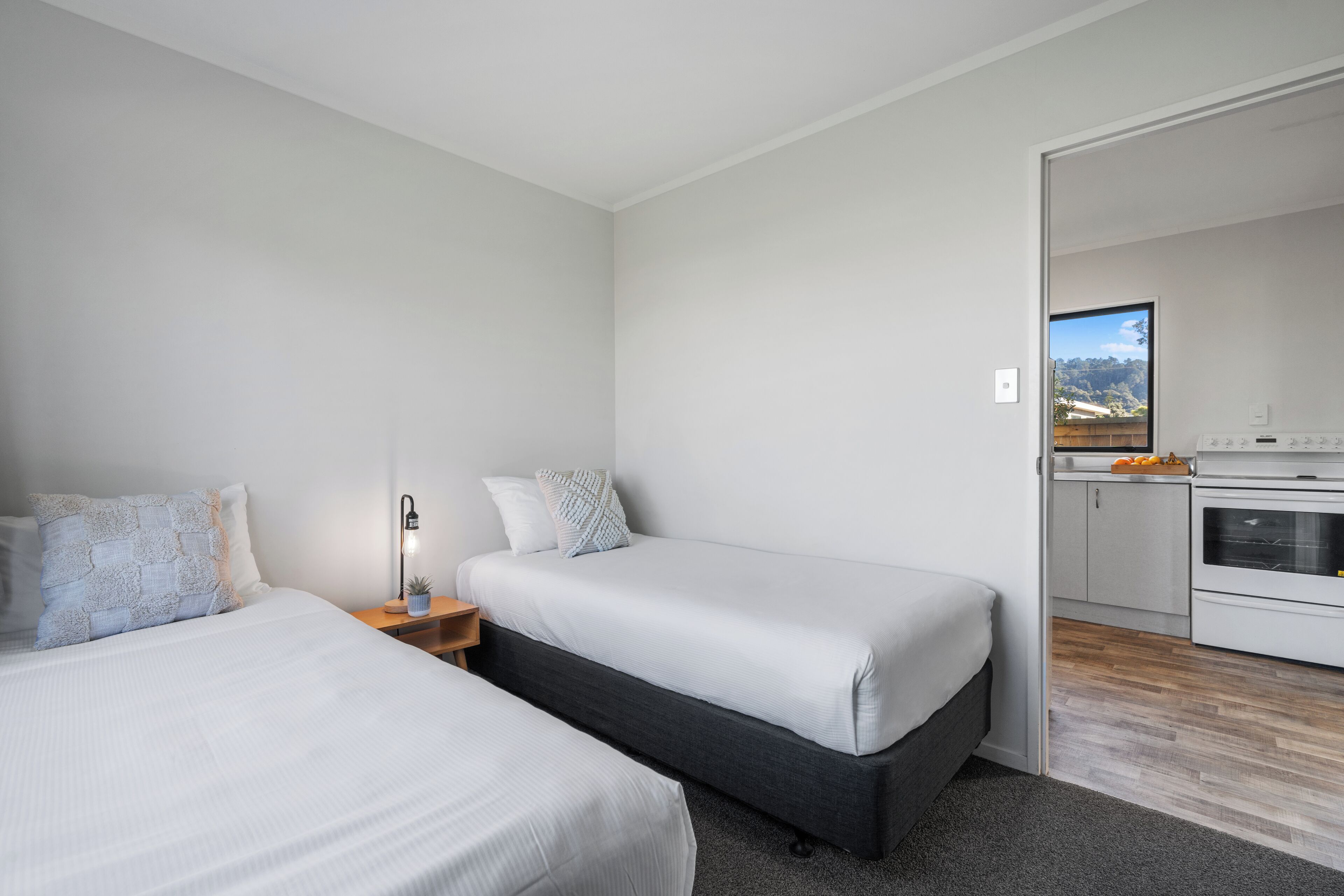 A modern bedroom with two beds, a bedside table with a lamp, and a view of a kitchen area.