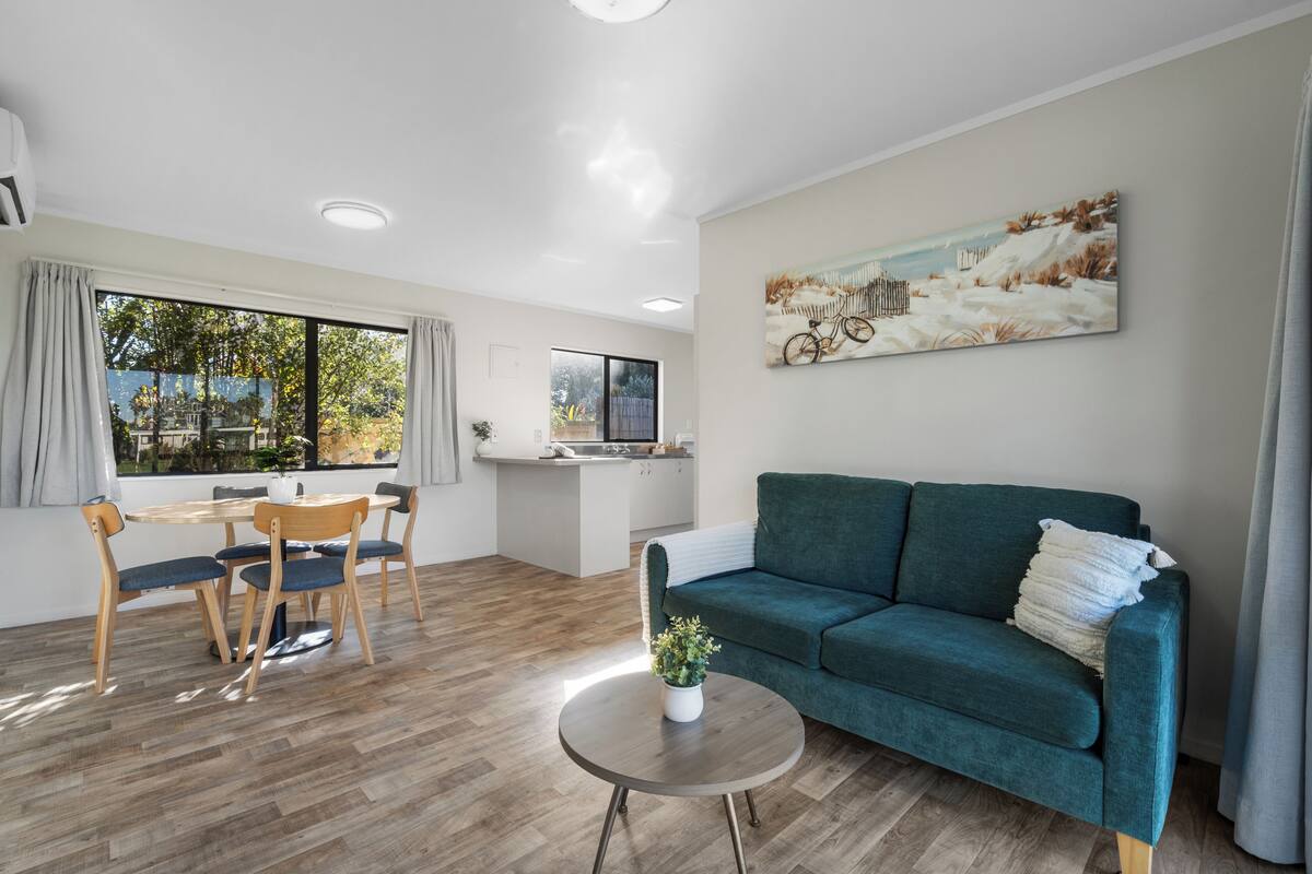 Una sala moderna con un sofá verde, una mesa de comedor de madera y sillas. Hay una zona de cocina con encimera y armarios blancos. Un cuadro de una bicicleta en un paisaje nevado cuelga de la pared.
