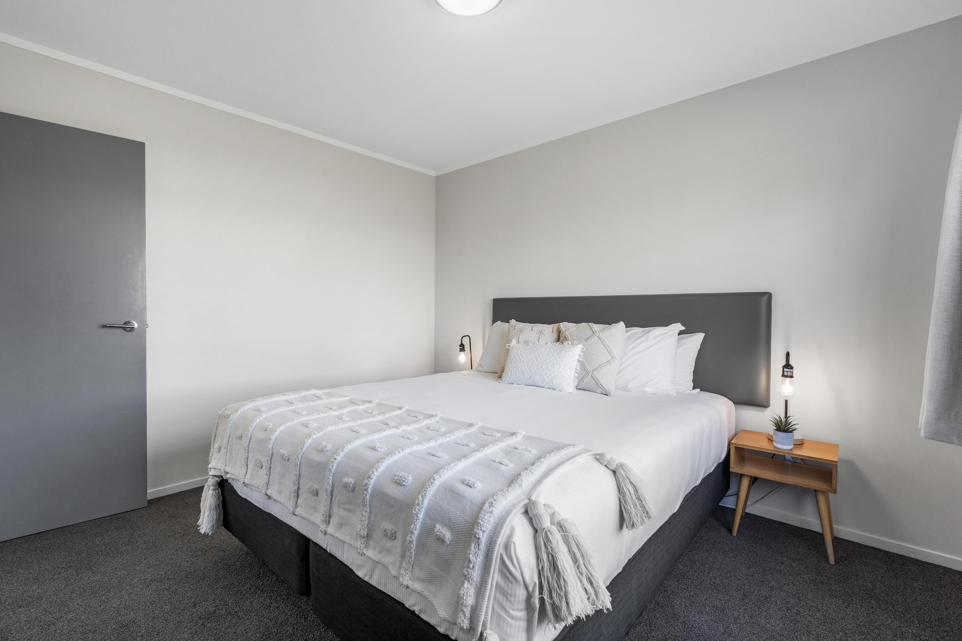 A bedroom with a large bed, a bedside table with a lamp, and a door.
