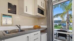 A modern kitchen with a built-in microwave, a sink, and a small refrigerator. There is a window with a view of a palm tree and a parking lot. - Tasman Holiday Parks – Beachaven (Waihi Beach)