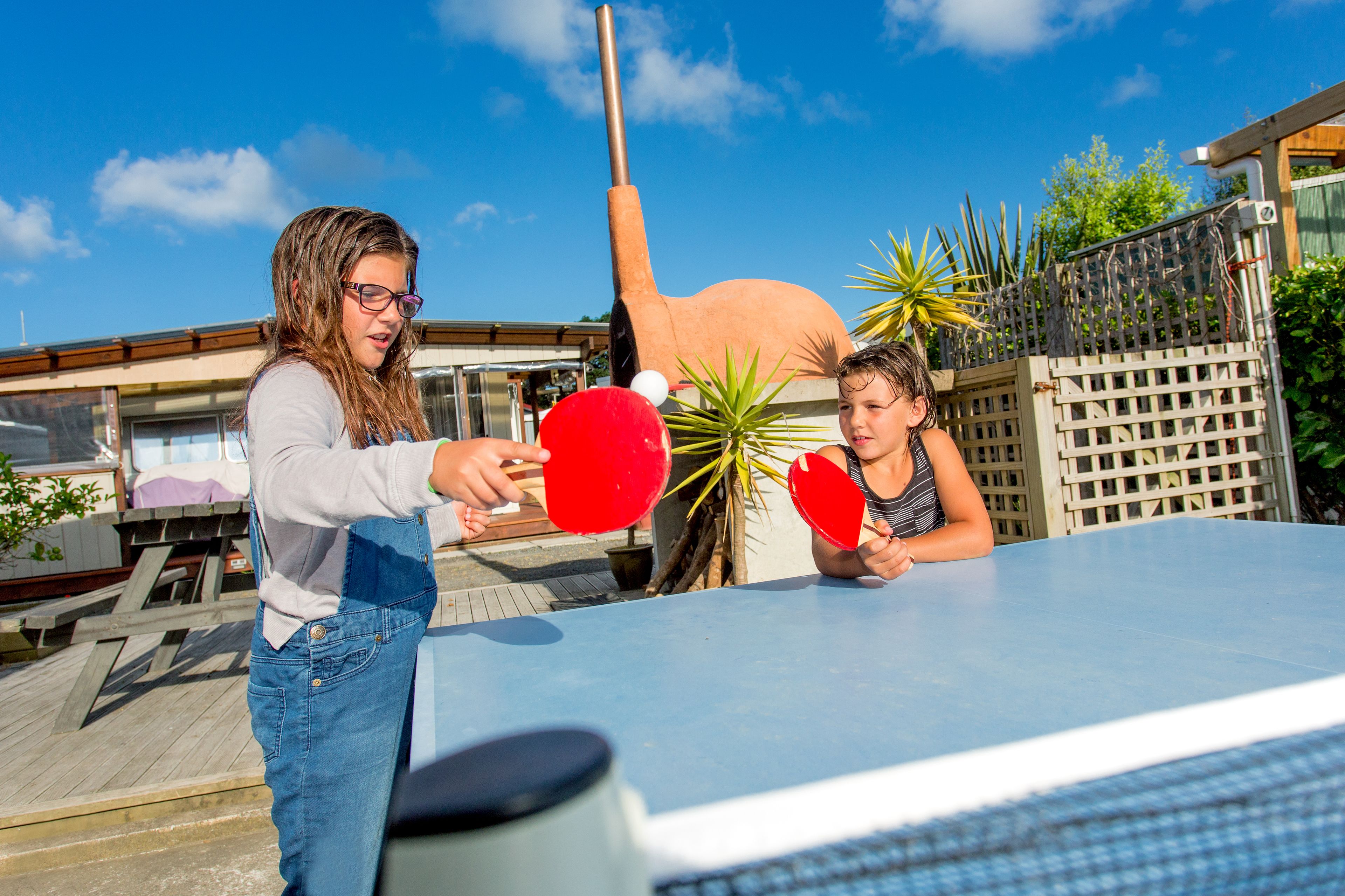 Dos personas jugando tenis de mesa al aire libre, con una mesa grande y una escultura llamativa al fondo.