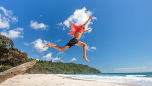 A beach scene with a person jumping in the air, a sandy beach, ocean waves, and a forested hill in the background. - Tasman Holiday Parks – Beachaven (Waihi Beach)