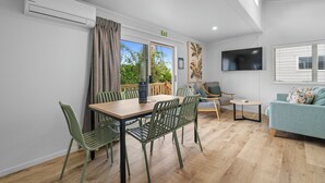 A modern living room with a dining area, a sofa, a TV, and a balcony with a view of trees. - Tasman Holiday Parks – Beachaven (Waihi Beach)