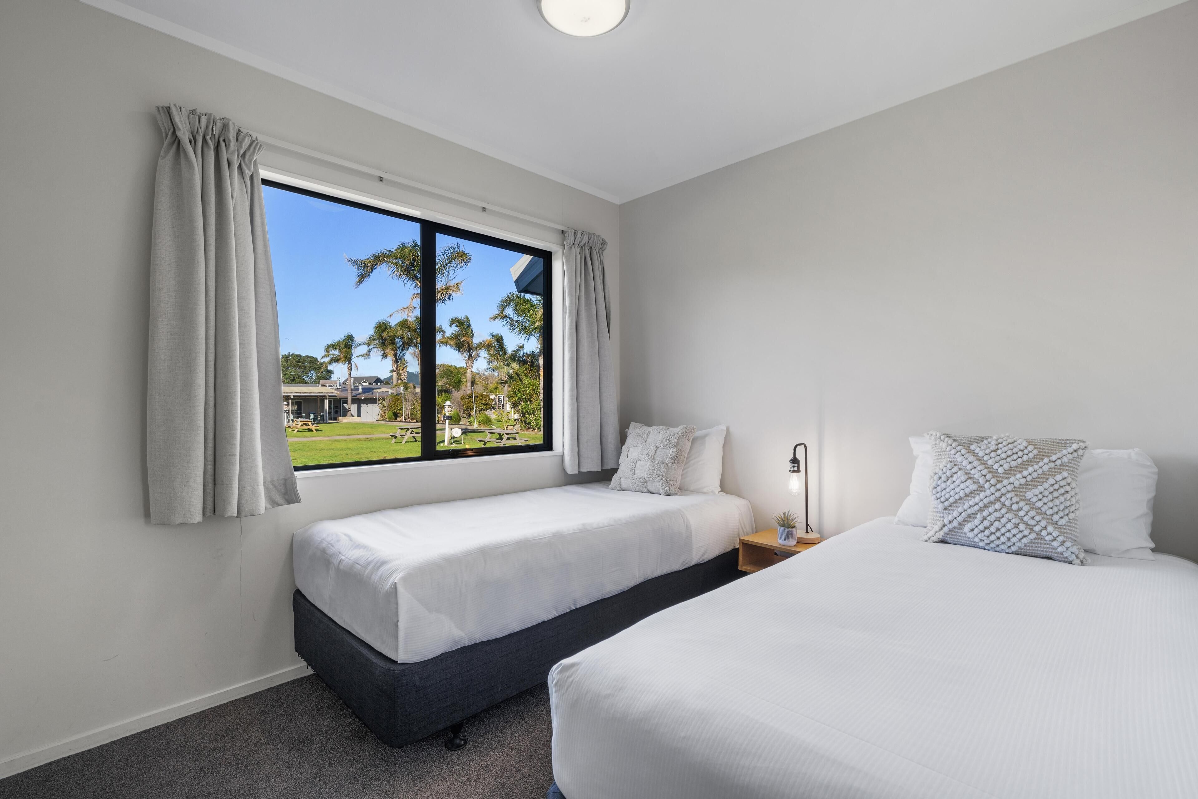 Habitación de hotel con dos camas, una ventana con cortinas, una mesita de noche con lámpara y vistas a palmeras y un edificio.