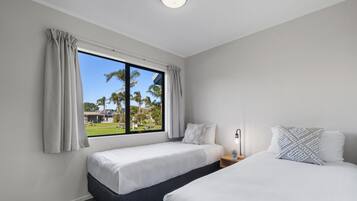 Habitación de hotel con dos camas, una ventana con cortinas, una mesita de noche con lámpara y vistas a palmeras y un edificio.