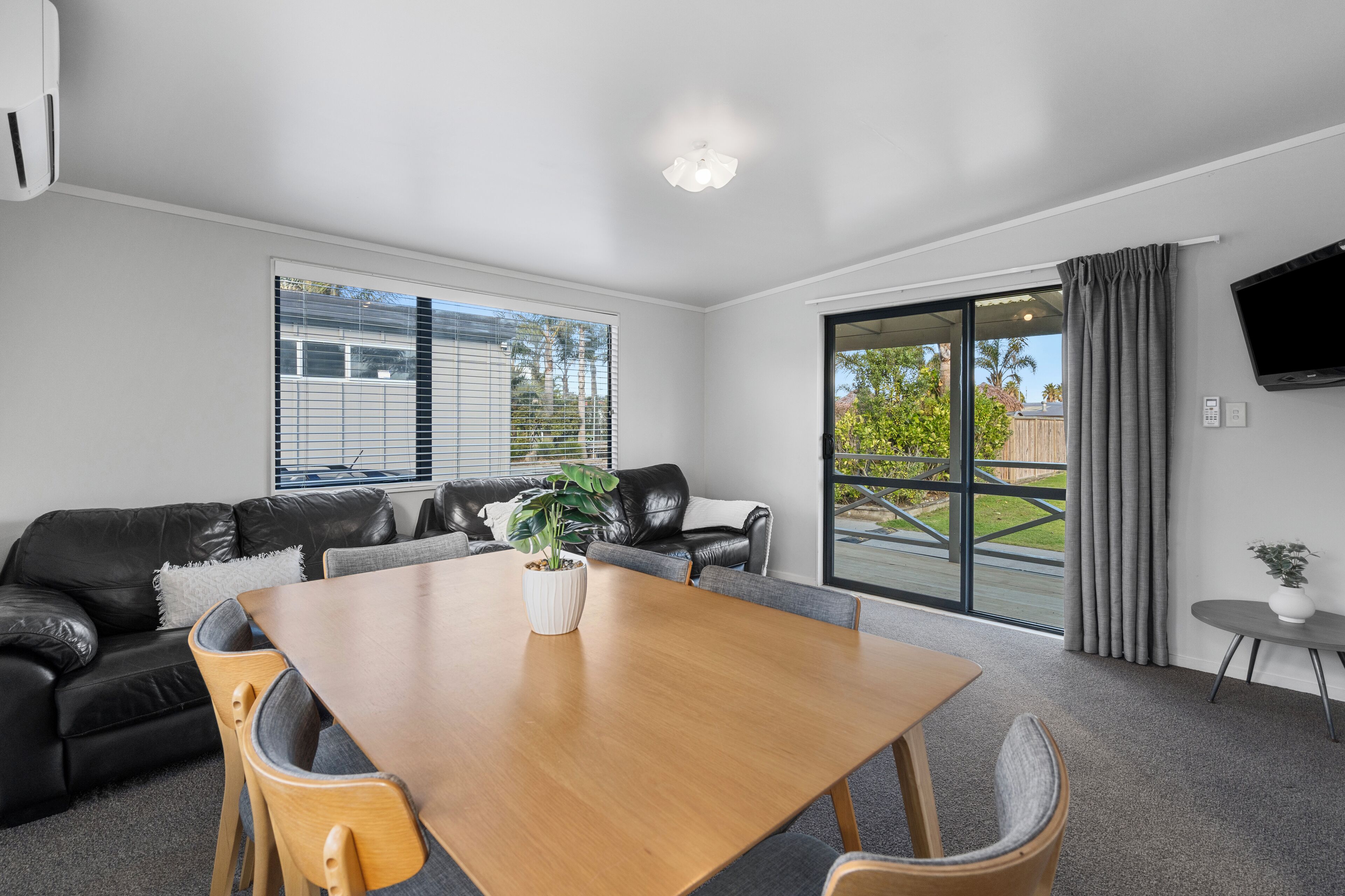 A modern living room with a wooden dining table, black leather sofa, and a large window with blinds.