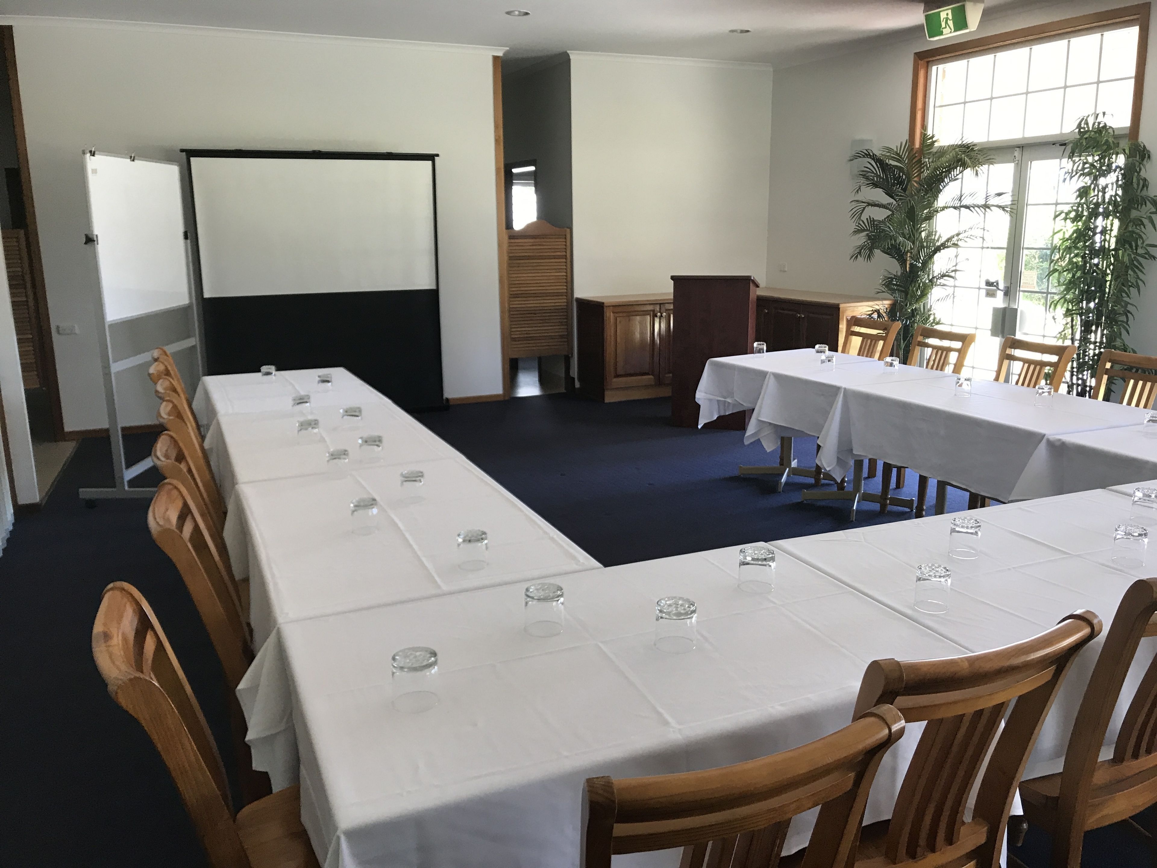 Un salón de conferencias con mesas largas, sillas, una pizarra y un amplio ventanal con plantas.