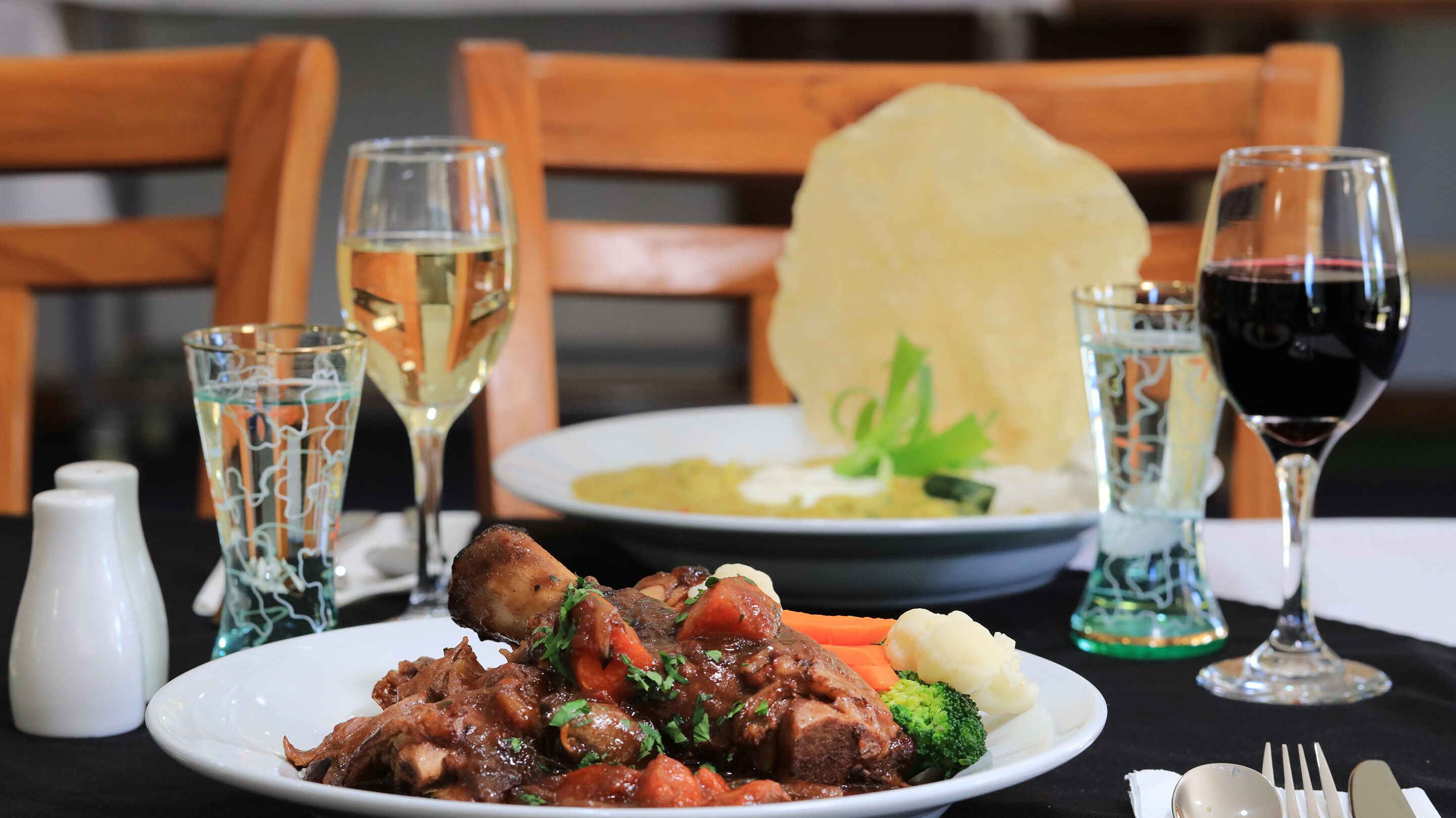 A dining table with a plate of food, two glasses of wine, and a salt and pepper shaker.